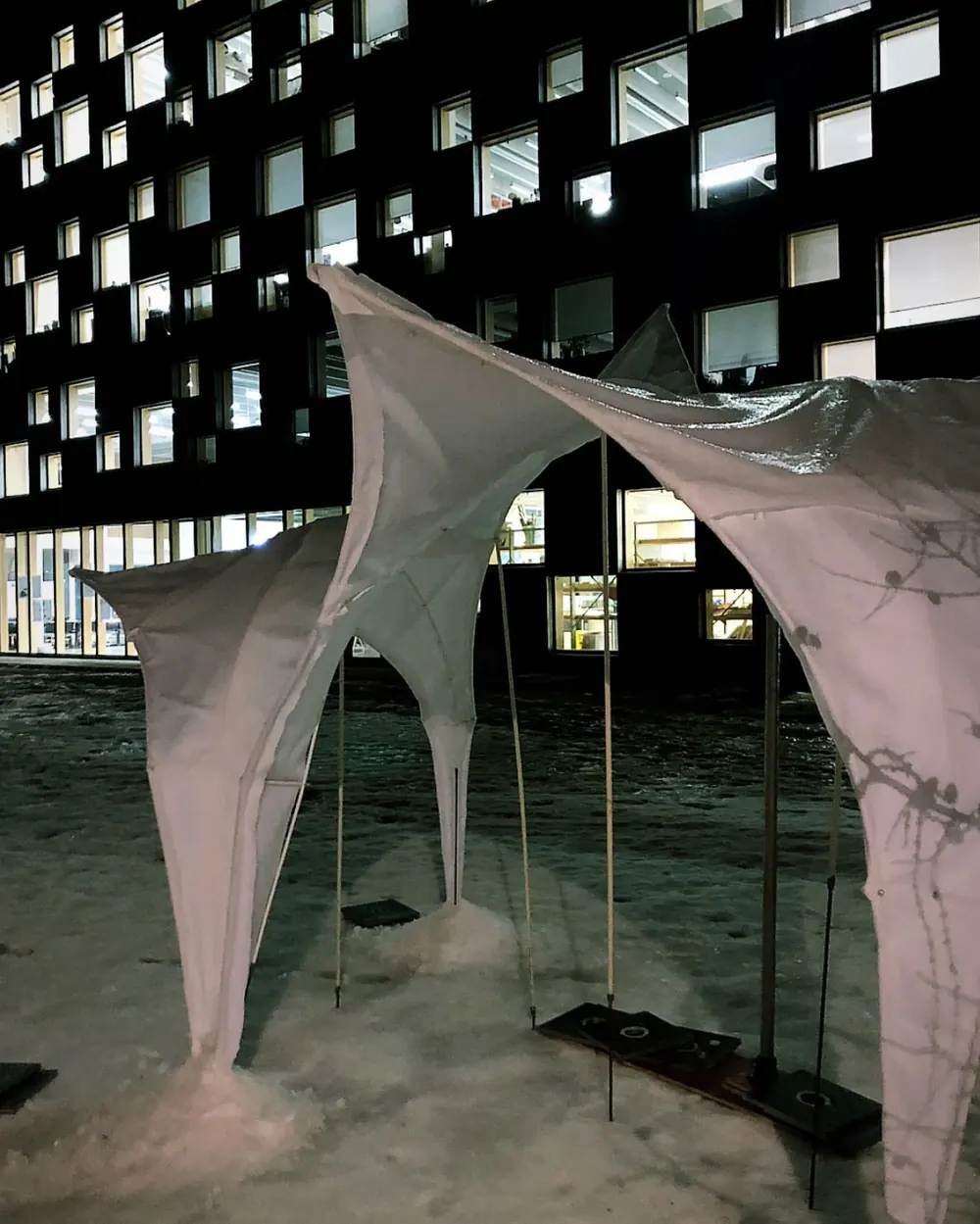 A white fabric minimal surface standing in the snow at night, in front of the lit school building