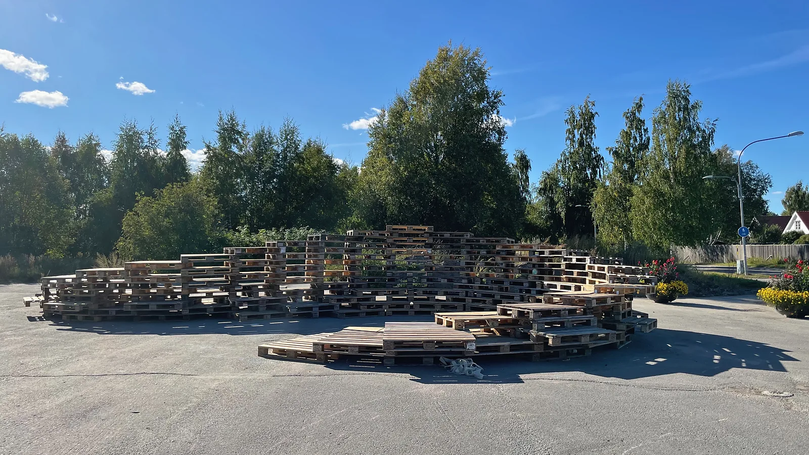The finished Pallet Parliament seen whole across the empty parking lot, its stacked tiers curving round under a blue sky